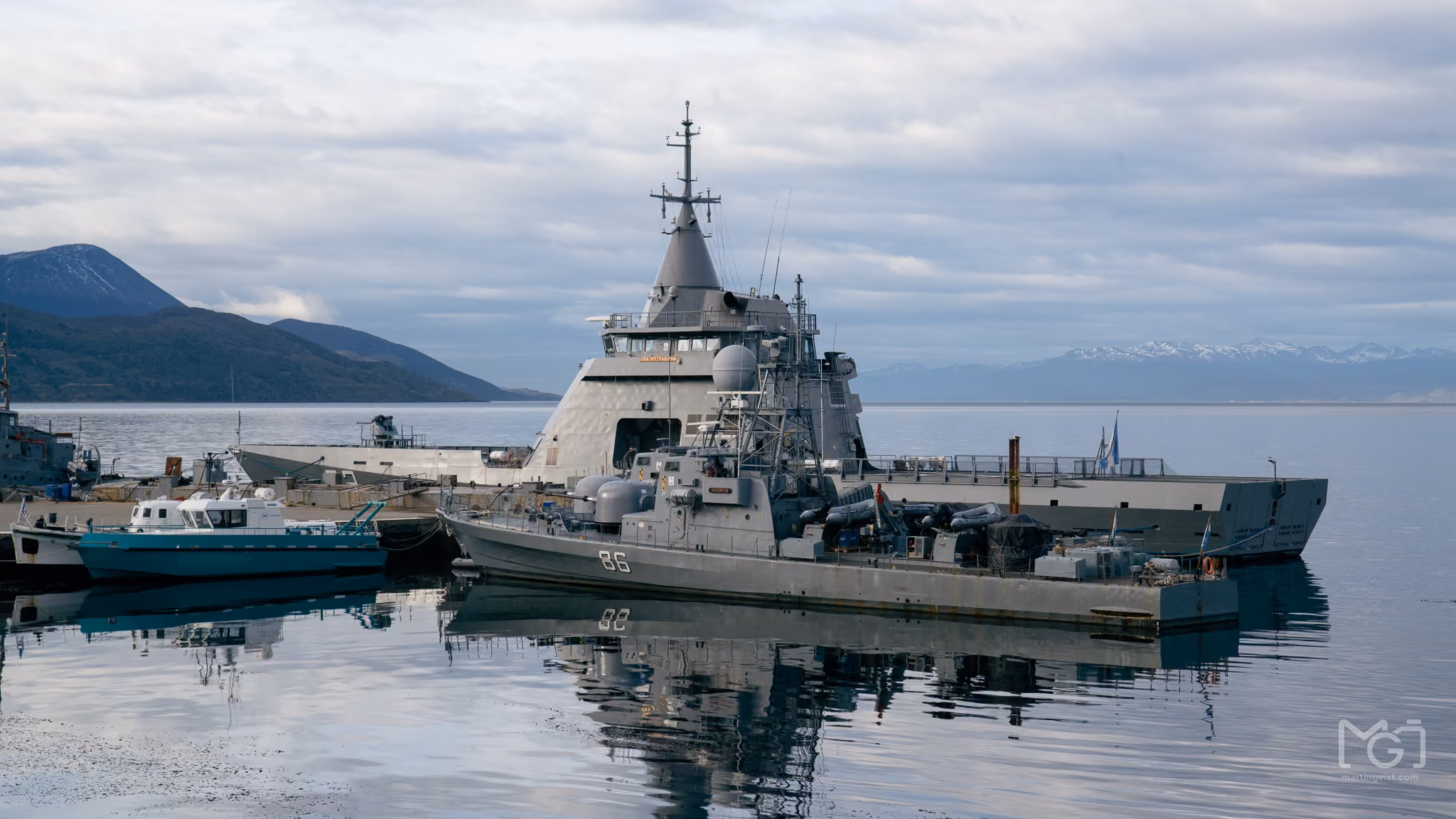 A grey naval ship moored in still water below mountains