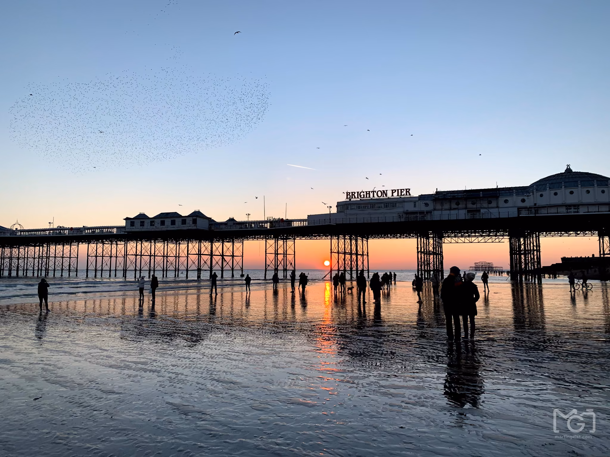 A pier at dusk with people on the wet sand below